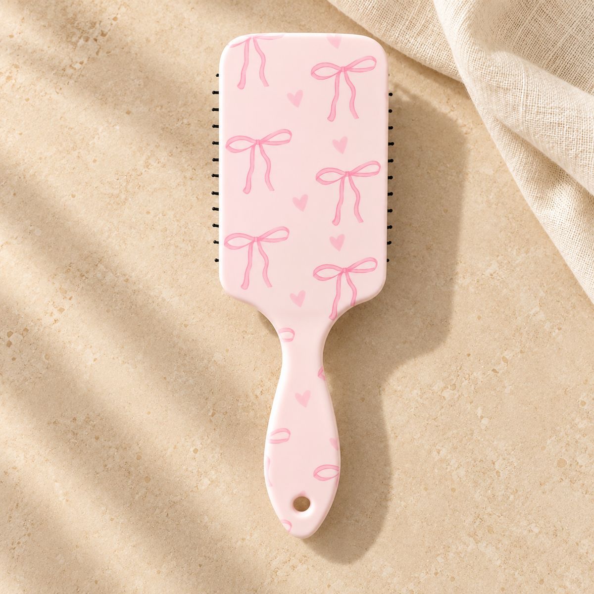 Back of a pale pink paddle hairbrush decorated with pink bows and hearts, with a hanging hole in the handle.