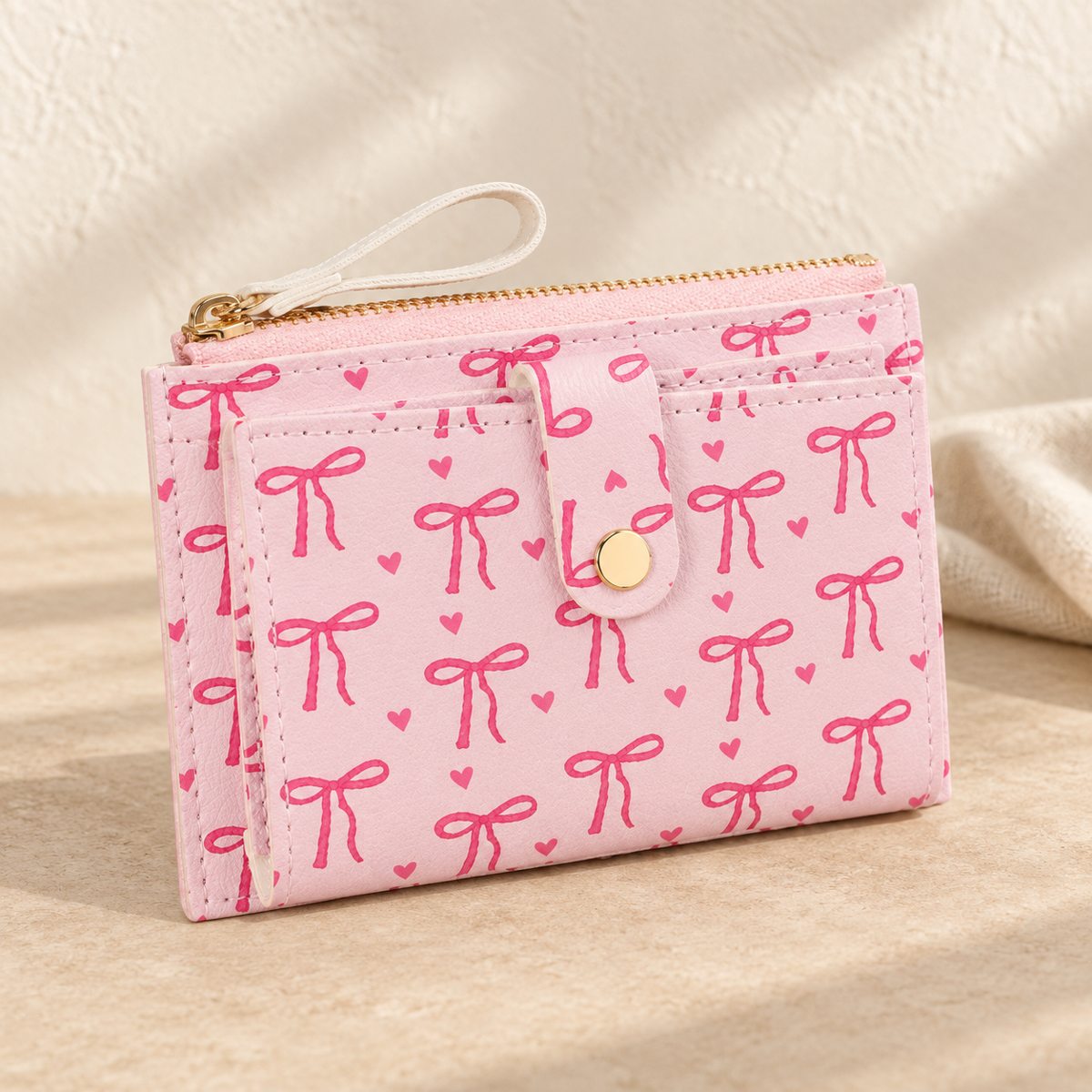Pale pink wallet with bow and heart print, gold-tone snap tab, zipped pocket and wrist strap.