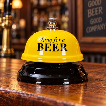 A novelty bar bell with a bright yellow dome top and black base, printed with the text 'Ring for a BEER' in black. It is set on a polished wooden bar counter with a blurred bar interior in the background.