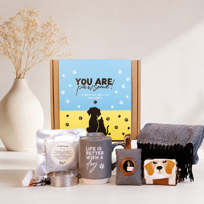 A curated pet gift set arranged on a white surface in front of a branded cardboard box. The set includes a gray ceramic mug with white text, a gray pouch, a metal bowl, a small gray pouch with a circular patch, a beagle-face novelty item, and a dark gray fringed textile.