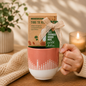 Hand holding a pink and white ceramic mug gift set featuring a heart-shaped aromatherapy eye pillow, decorative bow, and inspirational tag, styled in a cozy setting with a candle, knitted blanket, and indoor plant.

