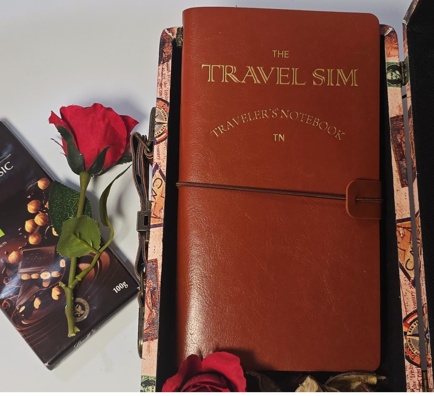 Vintage suitcase travel gift box containing a Lindt Swiss Classic chocolate bar, travel planner/sim book, and red artificial rose.