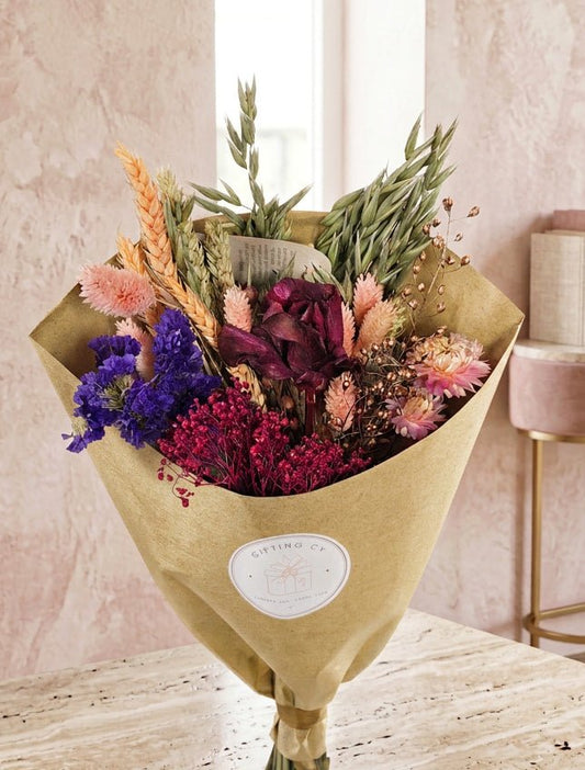 Elegant gift basket featuring a dried flower bouquet, decorative frame, ceramic vase, chocolate, tea, marshmallows, and a patterned mug, beautifully arranged in a woven basket with a pink ribbon against a soft neutral background.