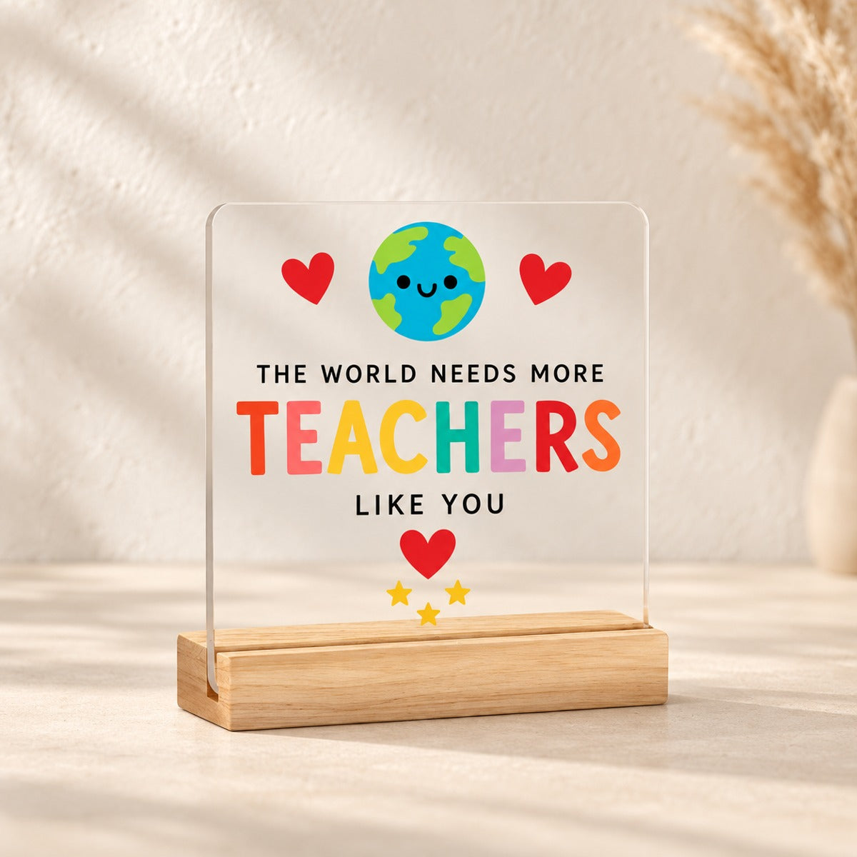Clear acrylic plaque reading The World Needs More Teachers Like You, with a smiling globe, red hearts and yellow stars on a wooden stand.