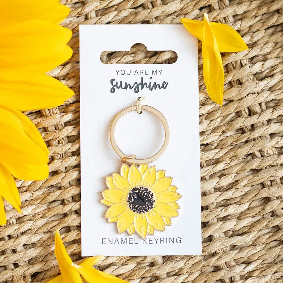 Yellow sunflower enamel keyring with “You are my sunshine” message on packaging, placed on a woven surface with sunflower petals.